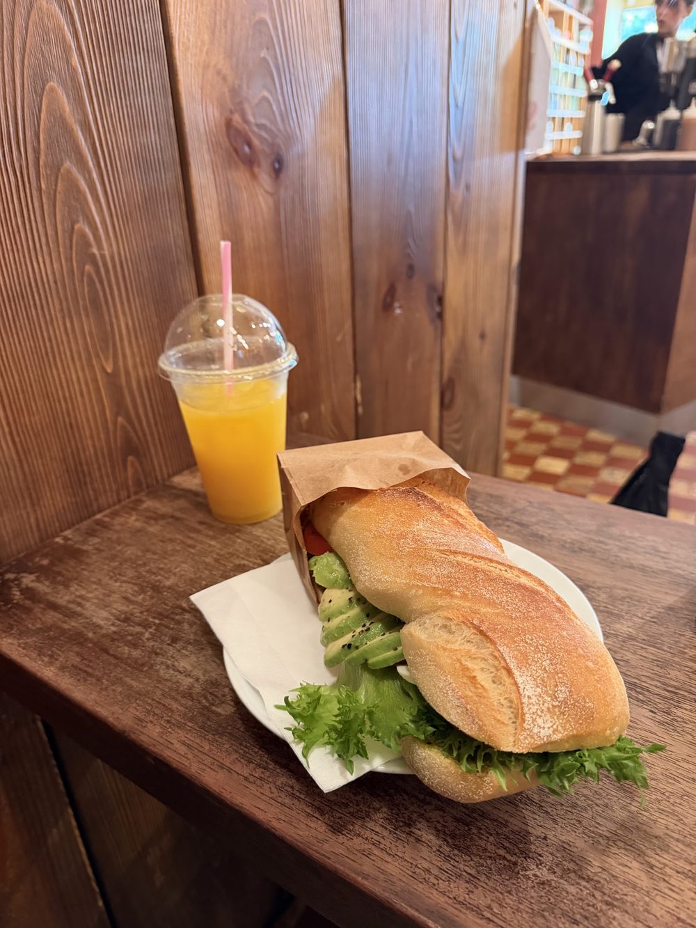 Baguett med avokado og salat, og et glass juice, på et trebord inne hos Evita